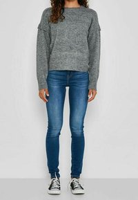 Personne portant un pull en tricot gris, un jean bleu ajusté et des baskets noires et blanches à lacets, debout devant un fond uni.