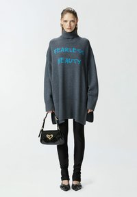 Sivi oversized pulover s natpisom "STRAH OSLIČENJE" u plavoj boji, rebrastim rubom. Upravljen s crnim tajicama i crnom patentnom torbom s zlatnim detaljem srca.