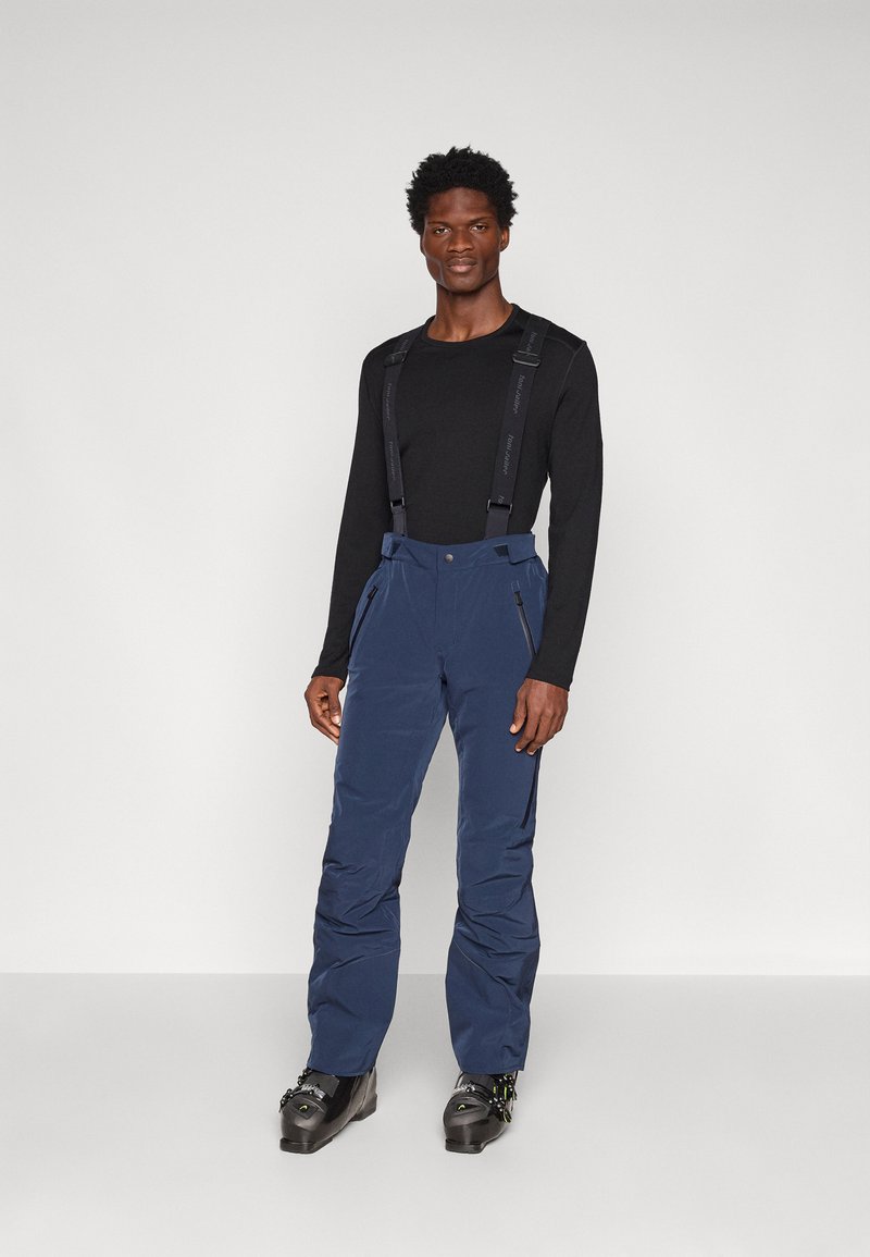 Navy waterdichte broek met verstelbare bretels, zakken met rits aan de voorkant, taps toelopende pijpen en versterkte zoom. Draag over een zwart overhemd met lange mouwen.