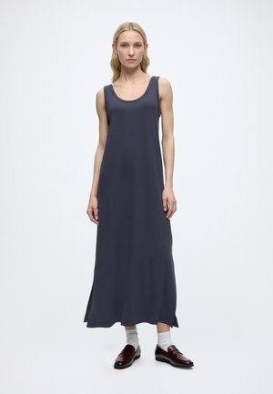 Γυναίκα με ξανθά μαλλιά που φοράει αμάνικο, μακρύ μέχρι τον αστράγαλο σκούρο γκρι φόρεμα, λευκές κάλτσες και σκουρό καφέ μοκασίνια, στέκεται μπροστά σε λευκό φόντο.