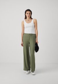 White sleeveless tank top paired with loose, olive green wide-leg pants. Model holding a black textured bag; wearing white sneakers.