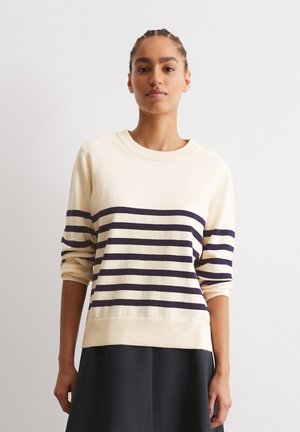 Maglione color crema con righe orizzontali blu navy, colletto rotondo e polsini arrotolati. Texture morbida e vestibilità comoda. Abbinato a una gonna scura.