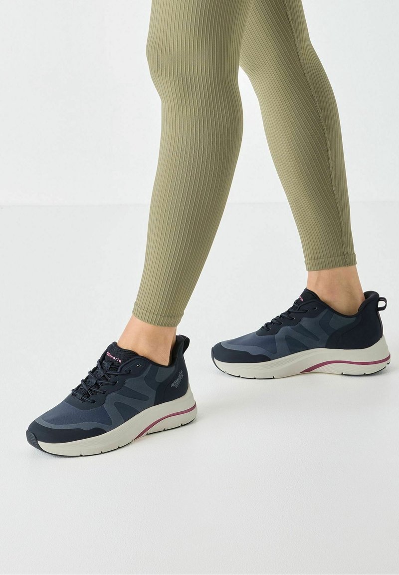 Jambes en leggings côtelés vert olive portant des baskets noires et gris foncé avec semelles blanches et touches roses sur un sol clair.