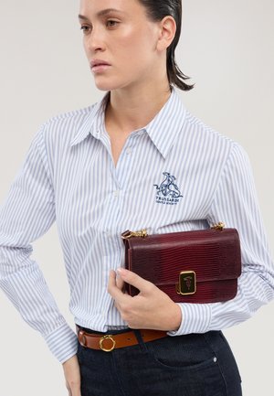 Vrouw draagt een wit-blauw gestreept shirt met een logo, en houdt een gestructureerde bordeauxrode handtas met gouden sluiting en kettingriem vast.