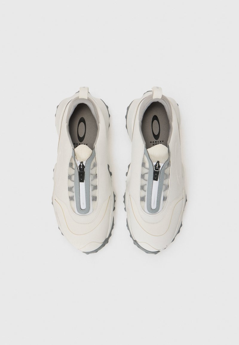 Beige slip-on shoes with a textured surface, grey accents, and a central zipper. The sole features rugged traction patterns.