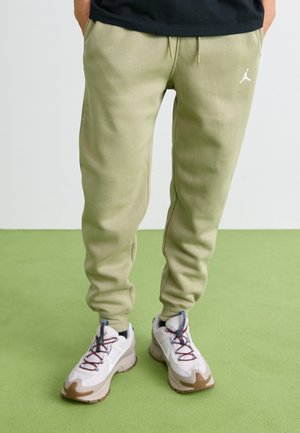 Person iført lysegrønne joggers med et hvidt logo og hvide sneakers med blå og brune detaljer, stående på et grønt gulv.