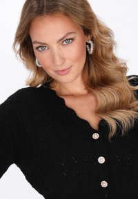 Schwarze Strickjacke mit strukturiertem Muster, V-Ausschnitt und fünf dekorativen Knöpfen mit perlenähnlichen Akzenten. Die Haare sind in lockeren Wellen gestylt.