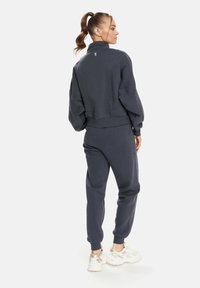 Dunkelgrauer Sweatshirt und passende Sweatpants aus weichem Stoff, mit hohem Kragen und elastischen Bündchen. Das Modell trägt Sneakers.