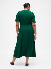 Woman with dark hair in a bun wearing a green midi dress with puffed sleeves, standing with back facing the camera on a white background.