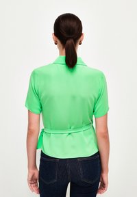 Grøn satin kortærmet bluse med krave, bæltestrop i taljen, glat tekstur og rygplissering, parret med mørke denim jeans.