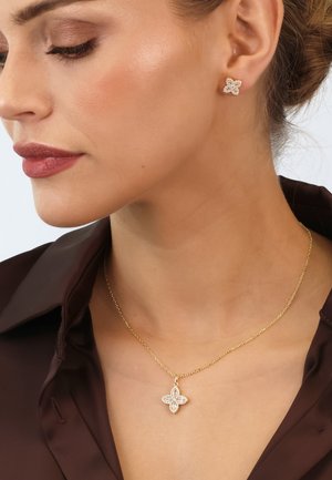 Goldkette mit einem floralen Anhänger, verziert mit kleinen Steinen. Das Modell trägt passende florale Steckerohrringe. Braunes Hemd.