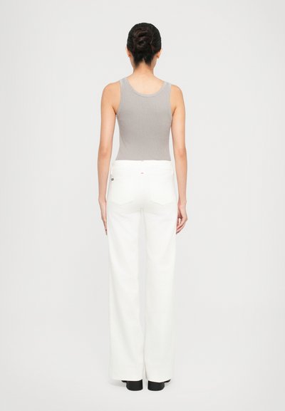 Femme aux cheveux bruns en chignon portant un haut sans manches gris, un pantalon large blanc et des chaussures noires, se tenant face à un fond blanc.