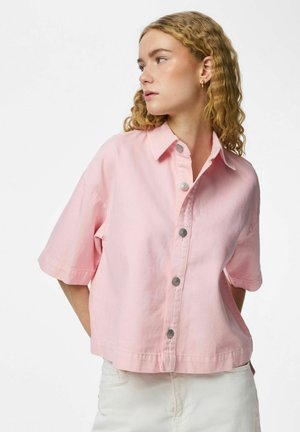 Mladá žena s vlnitými blond vlasmi, oblečená v voľnej ružovej košeli s krátkym rukávom a bielych nohaviciach, pozerá sa doľava.