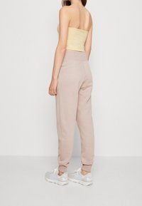 Kvinna stående i beige joggers, krämfärgad strapless topp och vit-grå sneakers mot en slät ljus bakgrund, sedd bakifrån.