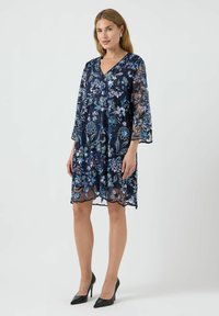 Vestido de encaje floral azul marino con mangas campana 3/4, que presenta un bordado intrincado en tonos azules y plateados, dobladillo con borde ondulado y escote en V.