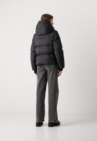 Veste doudoune noire avec une capuche à col haut, design matelassé et poignets côtelés, associée à un pantalon en denim gris ample et des chaussures noires imposantes.