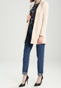 Cappotto beige su misura con colletto a punte sopra un top nero a fiori; abbinato a jeans blu con risvolti e tacchi alti neri.