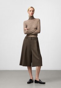 Jasnobrązowy sweter z prążkowanym golfem w połączeniu z ciemnozielonymi szerokimi spodniami culottes. Modelka ma na sobie czarne mokasyny. Gładkie tekstury; minimalistyczny design.