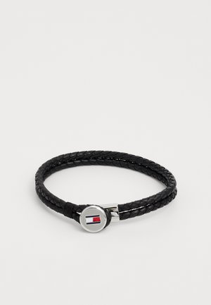 Schwarzes geflochtenes Lederarmband mit einem runden silbernen Verschluss, der mit einem rot, weiß und blau gestalteten Logo verziert ist. Glatte Textur und flexibles Design.