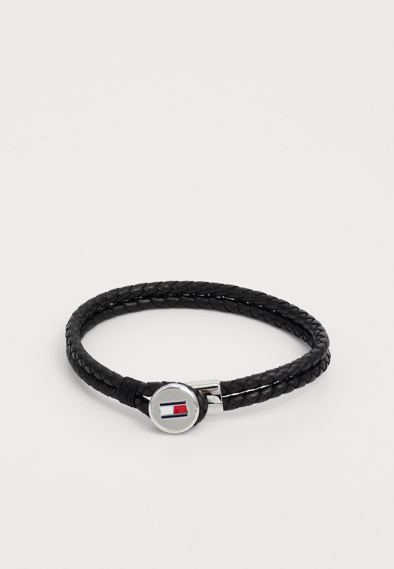 Must musträtikest punutud nahkkäevõru, millel on ümar hõbedane klamber koos punase, valge ja sinise logo detailiga. Sile tekstuur ja paindlik disain.