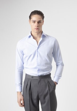 Joven con el cabello peinado hacia atrás, que lleva una camisa azul claro abotonada y metida dentro de unos pantalones grises plisados de cintura alta.