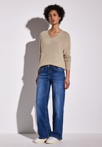 Béžový pletený svetr s výstřihem do V a dlouhými rukávy, doplněný volnými modrými džínami a bílými teniskami. Měkká textura, pohodlný střih a čistý design.