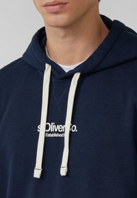 Uomo che indossa un hoodie blu navy con lacci bianchi e testo stampato bianco "s.Oliver co. Established 19" sul petto, visibile dal collo fino a metà torace.