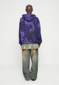 Felpa con cappuccio viola decorata con motivi floreali e animali; abbinata a una gonna con dettagli metallici sopra pantaloni a gamba larga grigi macchiati di vernice.