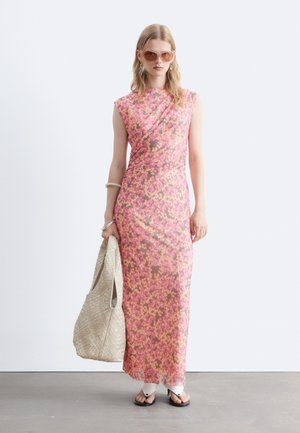 Donna che indossa un abito senza maniche, lungo fino ai piedi, con motivo floreale rosa e arancione, occhiali da sole, sandali bianchi, che tiene una grande borsa tote beige intrecciata.