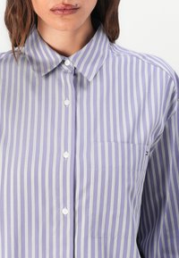 Camicia button-up a righe viola con accenti bianchi, caratterizzata da un colletto a punta, tasca frontale e texture del tessuto liscia.