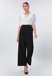 White loose blouse with flutter sleeves featuring silver embellishments, paired with high-waisted black wide-leg trousers. Black heels.