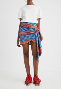 Kietaisuhame, jossa on sinisiä, oransseja ja punaisia geometrisiä kuvioita, yhdistetty valkoiseen lyhythihaiseen paitaan ja punaisiin hapsullisiin sandaaleihin.