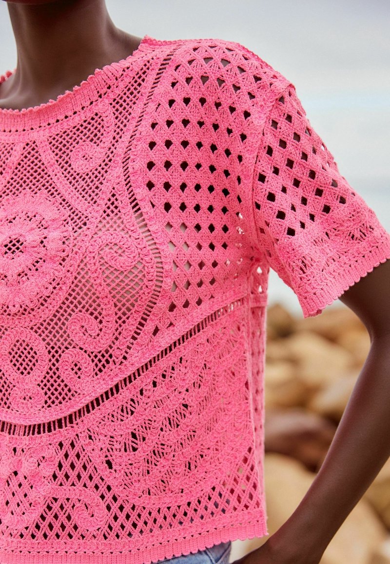 Różowy, ażurowy top z dzianiny, z krótkimi rękawami, złożony z misternych wzorów i falbaniastych krawędzi, prezentujący lekką i teksturowaną konstrukcję.