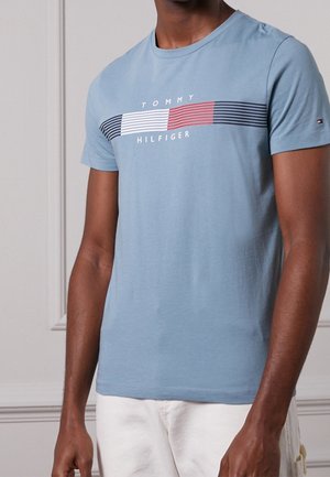 Mand iført en lyseblå Tommy Hilfiger T-shirt med korte ærmer og logo med røde, hvide og marineblå striber samt hvide bukser.