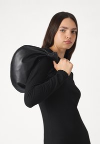 Black leather backpack with a soft texture, rounded shape, and stylish drawstring closure, being carried on the shoulder.