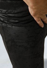 Zwarte neplederen broek met een textuurafwerking, voorzien van een getailleerd ontwerp en zichtbare stiksels. Een hand steunt op de tailleband.