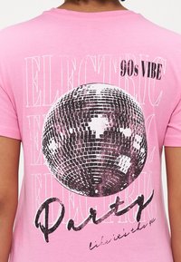Rosa T-skjorte med et stort svart-hvitt disco kule grafikk, fet tekst som sier "90s VIBE" og "PARTY," og en myk bomullstekstur.