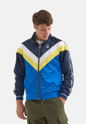 Bunte Sportjacke mit blauen, navyblauen, gelben und weißen Chevron-Mustern. Hergestellt aus glattem Stoff mit Reißverschluss vorne und Seitentaschen.