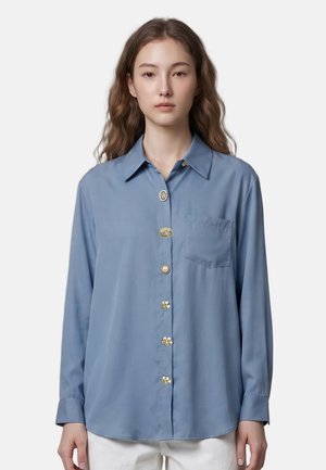 Femme portant une chemise bleue ample à boutons décoratifs uniques et une poche poitrine, associée à un pantalon blanc, debout devant un fond blanc.