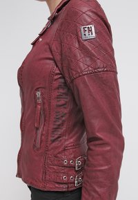 Bordeauxrode leren jas met een doorgestikte schouder, ritssluiting aan de voorkant en zijgespen. Voorzien van een logopatch op de mouw en een gestructureerd oppervlak.