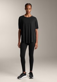 Musta lyhythihainen toppi pyöreällä kaula-aukolla, yhdistettynä mustiin legginseihin, joissa on rakenteellisia yksityiskohtia, sekä mustiin urheilukenkäpariin, joissa on valkoiset pohjat.