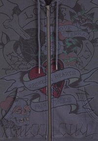 Felpa grigia con zip caratterizzata da un mix di design stampati, tra cui un cuore rosso con embellishments e banner di testo, realizzata in un tessuto testurizzato.