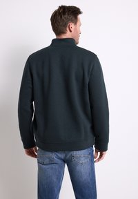 Uomo che indossa un pullover strutturato verde scuro e jeans blu, di spalle su uno sfondo chiaro e neutro.