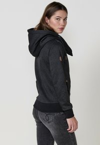 Felpa nera a motivo con un grande cappuccio, polsini e orlo a coste, e accenti marroni sulle spalle. Abbinata a jeans grigi.