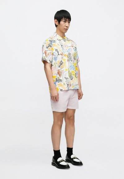 Κοντομάνικο, πολυχρωμο πουκάμισο με τυπωμένα αφηρημένα σχέδια· συνδυασμένο με ανοιχτό ροζ σορτς και μαύρα μοκασίνια με λευκές λεπτομέρειες.