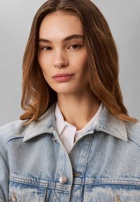 Jeune femme aux cheveux châtain clair ondulés portant une veste en jean bleu clair par-dessus une chemise blanche à col, fond gris neutre.