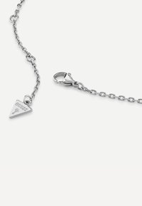 Bracelet en chaîne argentée avec un charme triangulaire portant le texte "GUESS" et une fermeture à mousqueton. Texture lisse et finition polie.