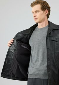 Zwarte leren jas met een knoopsluiting, binnenzak en een gladde textuur, gedragen over een grijs vest. Heeft een kraagontwerp.