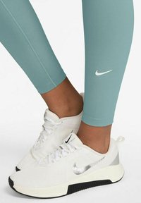 Leggings Nike di colore verde menta, con un design aderente e logo bianco a contrasto. Abbinati a sneakers bianche con accenti argento e suola nera.