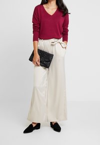 Burgunderfarbener V-Ausschnitt-Pullover, cremefarbene weit geschnittene Hose mit einer Schleife, schwarze strukturierte Clutch und schwarze Wildleder-Loafer.
